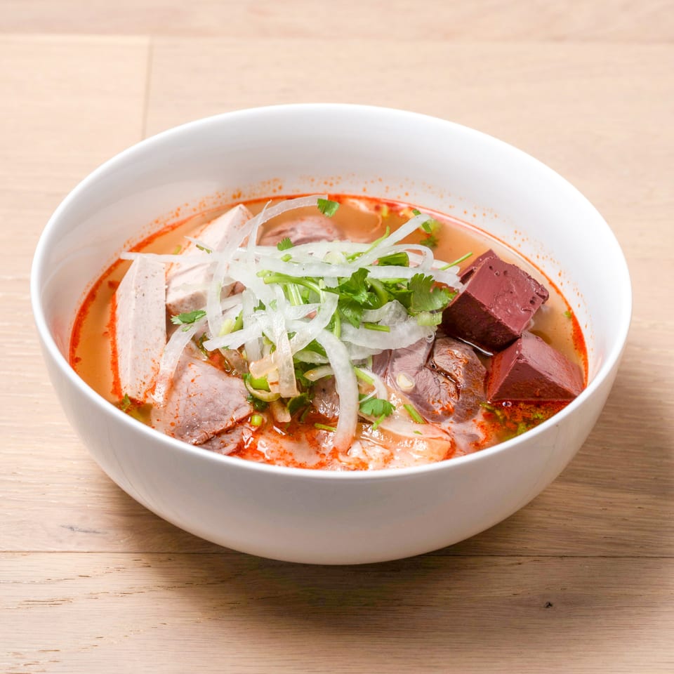 A white bowl of Bun Bo Hue, a spicy Vietnamese noodle soup with beef, pork, and blood cubes.
