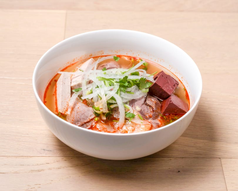 A white bowl of Bun Bo Hue, a spicy Vietnamese noodle soup with beef, pork, and blood cubes.