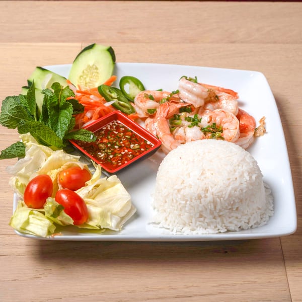 A white plate with shrimp, rice, fresh vegetables, and a red dipping sauce on a wooden table.