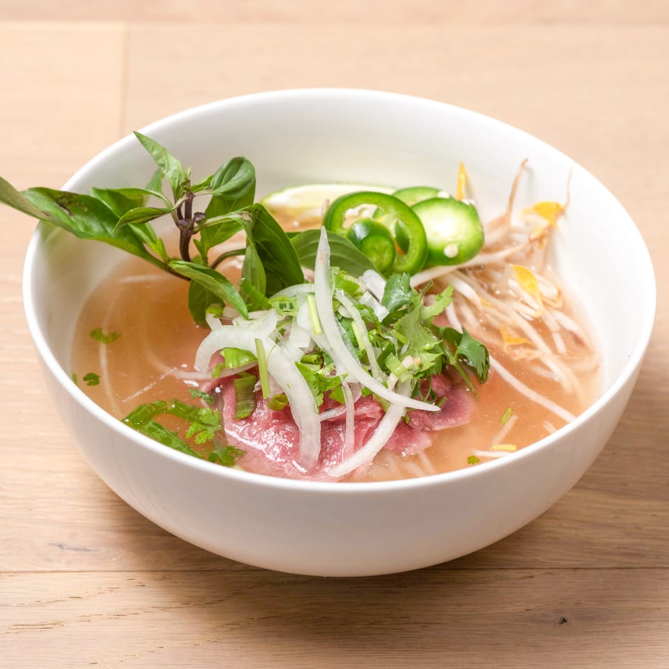A white bowl of pho with rare beef, bean sprouts, basil, jalapeños, and lime.