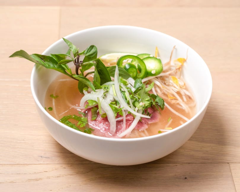 A white bowl of pho with rare beef, bean sprouts, basil, jalapeños, and lime.