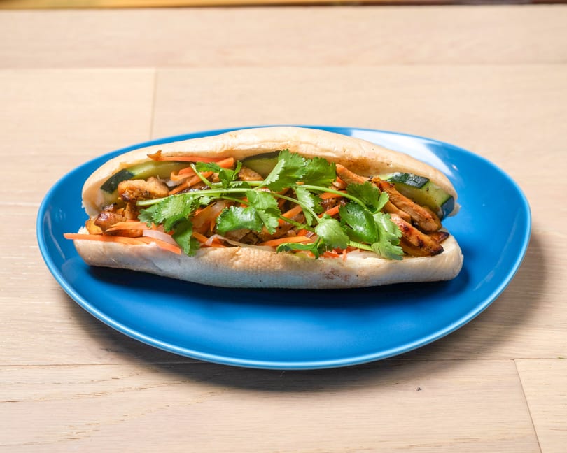 A banh mi sandwich with grilled chicken, pickled carrots, cucumbers, and cilantro on a blue plate.